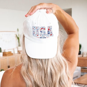 May include: A white baseball cap with the word "USA" in blue letters filled with a floral pattern of red and blue flowers. The text "EST. 1776" is below the word "USA".