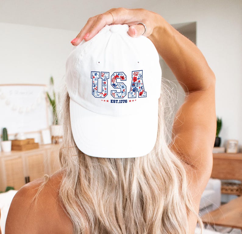 May include: A white baseball cap with the word "USA" in blue letters filled with a floral pattern of red and blue flowers. The text "EST. 1776" is below the word "USA".