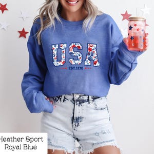 May include: Royal blue sweatshirt with the word "USA" in a floral design, with "EST.1776" below. The sweatshirt is paired with denim shorts. A glass with a straw and red and blue star design is held in the hand.