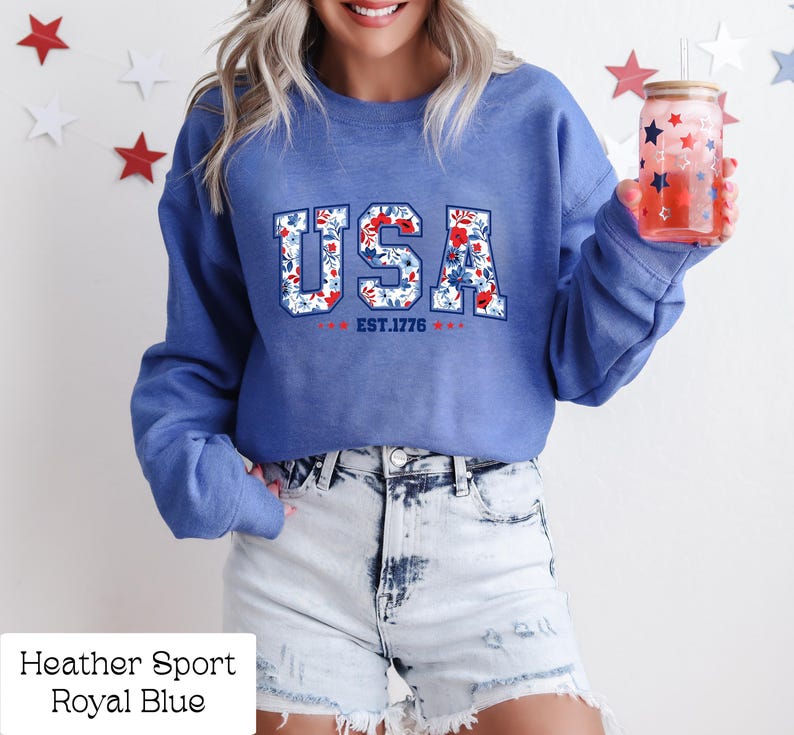 May include: Royal blue sweatshirt with the word "USA" in a floral design, with "EST.1776" below. The sweatshirt is paired with denim shorts. A glass with a straw and red and blue star design is held in the hand.