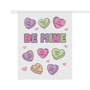 May include: A white garden flag with the words "BE MINE" in purple and green. The flag features heart-shaped candies with phrases like "Free Press," "Rule of Law," and "Due Process."