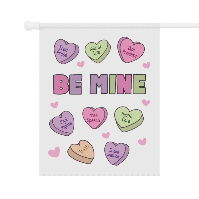 May include: A white garden flag with the words "BE MINE" in purple and green. The flag features heart-shaped candies with phrases like "Free Press," "Rule of Law," and "Due Process."