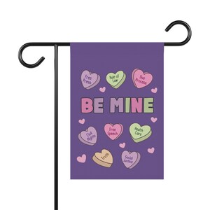 May include: A purple garden flag with the words "BE MINE" and heart-shaped candies with phrases like "Free Press" and "Social Justice." The flag is mounted on a black metal stand.