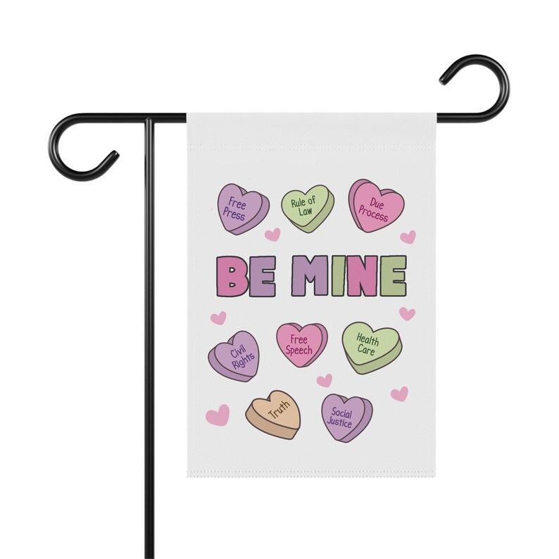 May include: A white garden flag with the words "BE MINE" in purple and green, surrounded by heart-shaped candies with phrases like "Free Press" and "Civil Rights." The flag is mounted on a black metal stand.