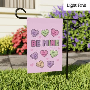 May include: A light pink garden flag with the words "BE MINE" in large letters. The flag features heart-shaped candies with phrases like "Free Press" and "Civil Rights". The flag is displayed on a black metal stand in a garden.