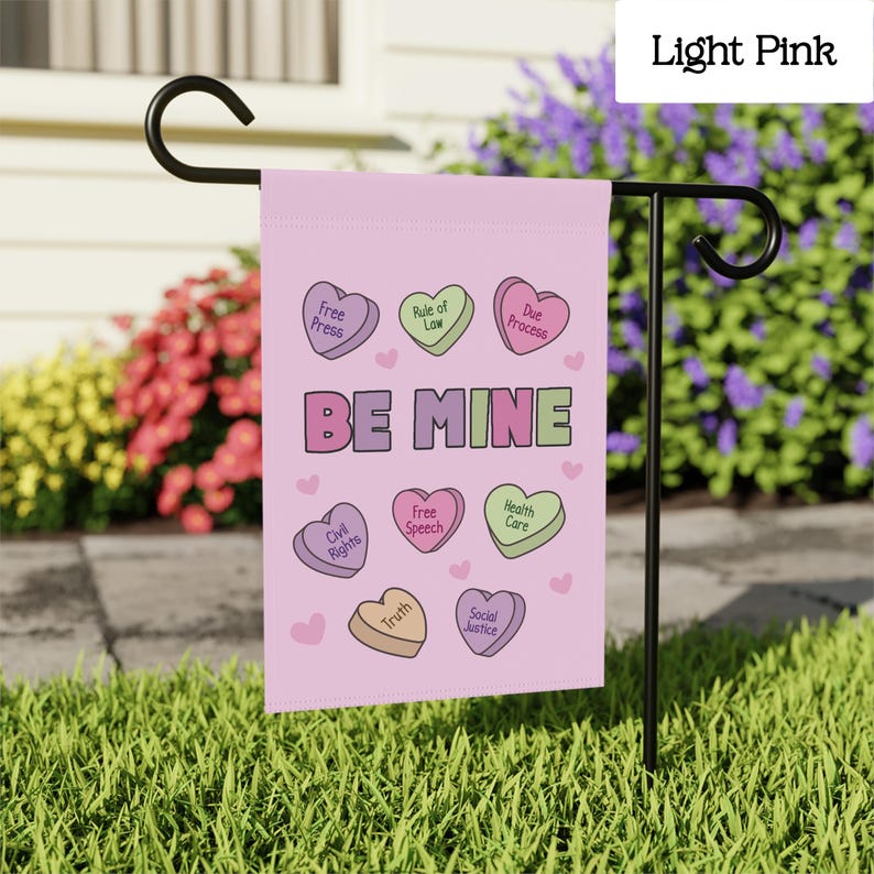 May include: A light pink garden flag with the words "BE MINE" in large letters. The flag features heart-shaped candies with phrases like "Free Press" and "Civil Rights". The flag is displayed on a black metal stand in a garden.