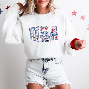 May include: White sweatshirt with the word "USA" in blue letters filled with a floral pattern of red and blue flowers. The text "EST.1776" is below the word "USA". The sweatshirt is paired with denim shorts.