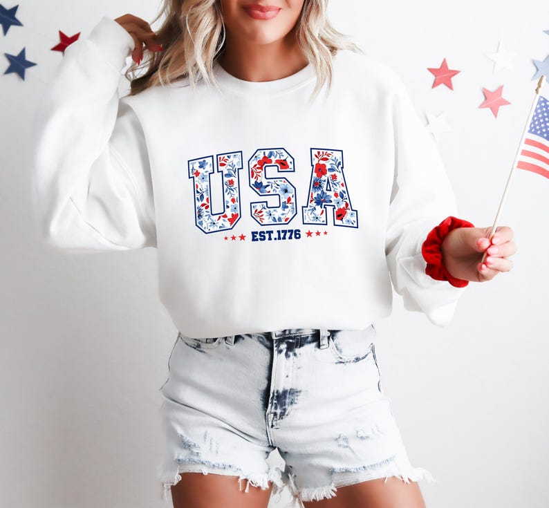 May include: White sweatshirt with the word "USA" in blue letters filled with a floral pattern of red and blue flowers. The text "EST.1776" is below the word "USA". The sweatshirt is paired with denim shorts.