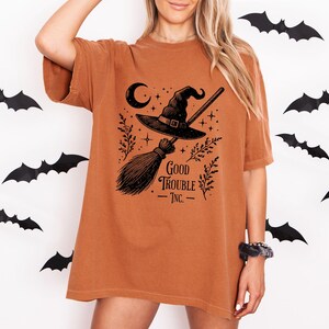 May include: A burnt orange short-sleeve t-shirt featuring a black graphic print of a witch hat, a broomstick, a crescent moon, stars, and leafy sprigs. The text Good Trouble Inc. is printed in black below the broomstick illustration.