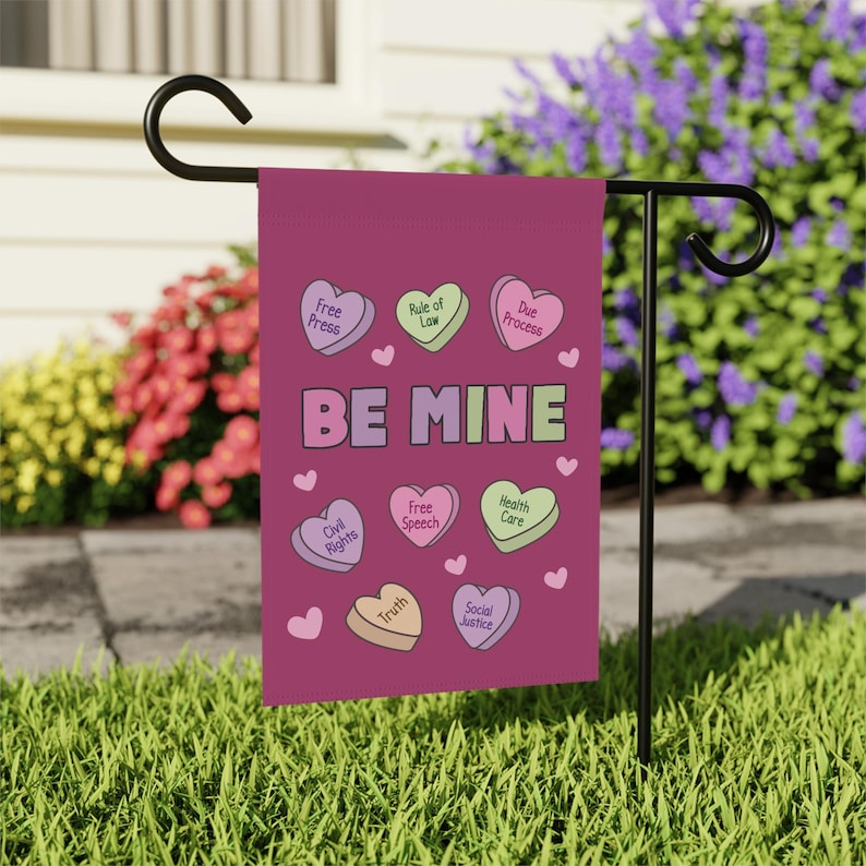 May include: A burgundy garden flag with the words "BE MINE" in pastel colors. The flag features heart-shaped candies with phrases like "Free Press," "Civil Rights," and "Social Justice." The flag is displayed on a black metal stand in a garden setting.