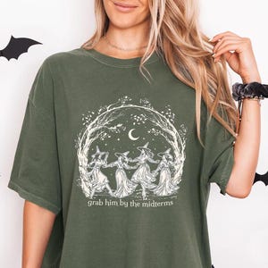 May include: An olive green short-sleeve t-shirt featuring a white graphic print of four figures in pointed hats dancing in a circle under a crescent moon and trees. The text "grab him by the midterms" is printed in white below the illustration.