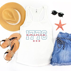 May include: A white vest top with the text "1776 america" in blue and pink, with three pink stars. The image also includes a straw hat, sunglasses, denim shorts, sandals, and a starfish.