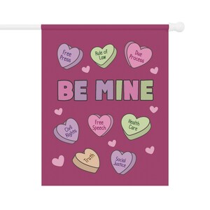 May include: A Valentine's Day themed garden flag with a dark pink background. The flag features heart-shaped candies with phrases like "Free Press" and "Be Mine". Small pink hearts are scattered around the design.
