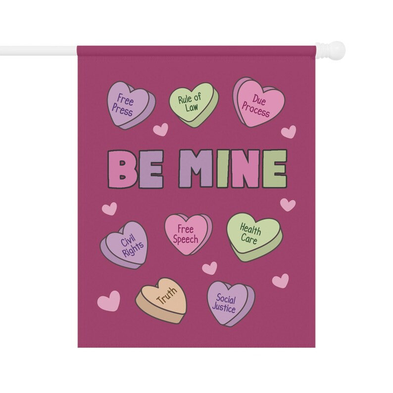 May include: A Valentine's Day themed garden flag with a dark pink background. The flag features heart-shaped candies with phrases like "Free Press" and "Be Mine". Small pink hearts are scattered around the design.