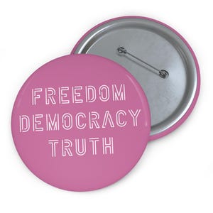 Op de afbeelding: Twee ronde roze buttons met de woorden "FREEDOM DEMOCRACY TRUTH" in witte, schreefloze letters. Eén button ligt voor de andere en toont de zilveren speldrug.