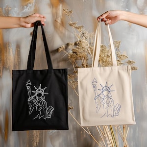 May include: Two tote bags, one black and one beige, each featuring a white or blue outline of the Statue of Liberty. The bags have long handles and are held up against a neutral background.