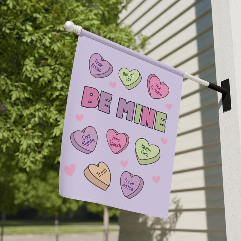 May include: A light purple flag with the words "BE MINE" in large letters. The flag features heart-shaped candies with phrases like "Free Press," "Civil Rights," and "Health Care." Small pink hearts are scattered around the design.