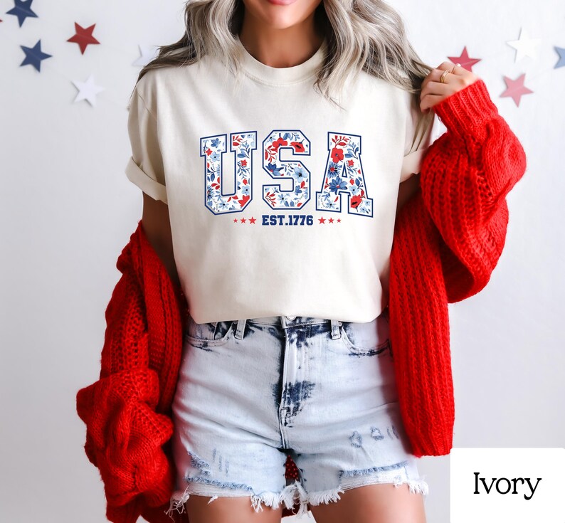 May include: Ivory-colored t-shirt with the word "USA" in a floral design, featuring red and blue flowers. The text "EST. 1776" is below the word "USA". The person is wearing denim shorts and a red sweater.