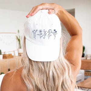 May include: White baseball cap with a blue embroidered design of five flowers with stems and leaves. The flowers are stylized and have a simple design.