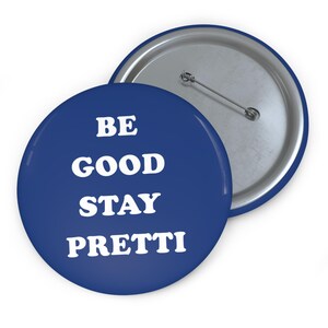 Pode incluir: Dois botões redondos. O botão frontal é azul escuro com o texto branco "BE GOOD STAY PRETTI". A parte de trás do botão é prateada com um alfinete. O segundo botão é parcialmente visível.