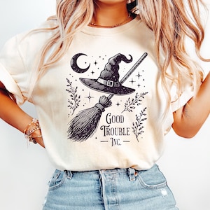 May include: Cream-colored t-shirt featuring a black and white graphic of a witch's hat, broom, and crescent moon. The text "Good Trouble Inc." is printed below the graphic. The shirt is made of a soft material.