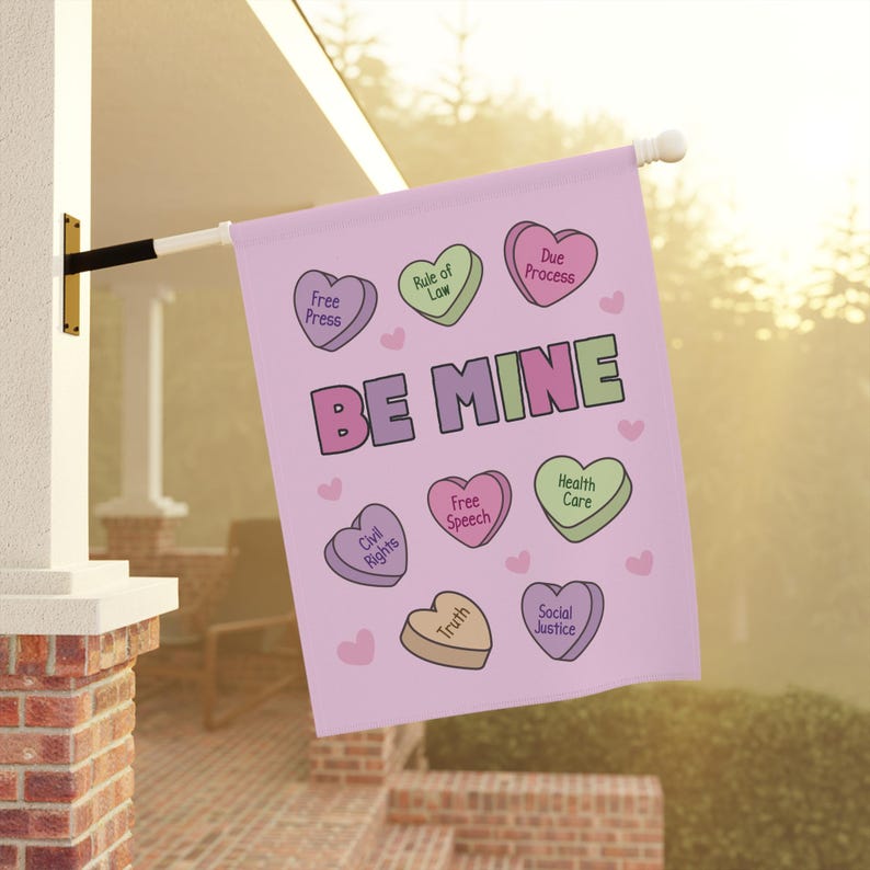 May include: A pink flag with the words "BE MINE" and heart-shaped candies with phrases like "Free Press" and "Social Justice." The flag is hanging outside on a white pole.