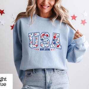 May include: Light blue sweatshirt with the word "USA" in a floral design, with the text "EST. 1776" below. The sweatshirt is worn by a person. The background has star decorations.