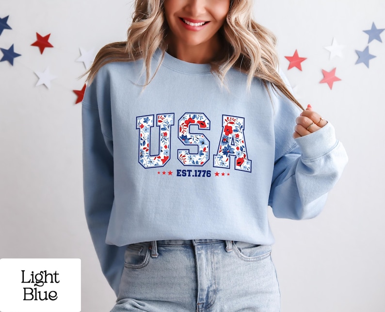 May include: Light blue sweatshirt with the word "USA" in a floral design, with the text "EST. 1776" below. The sweatshirt is worn by a person. The background has star decorations.
