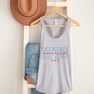 May include: A heather grey vest top with the text "1776 America" in red and blue. The top is displayed on a wooden hanger, with a brown hat and blue jeans in the background.