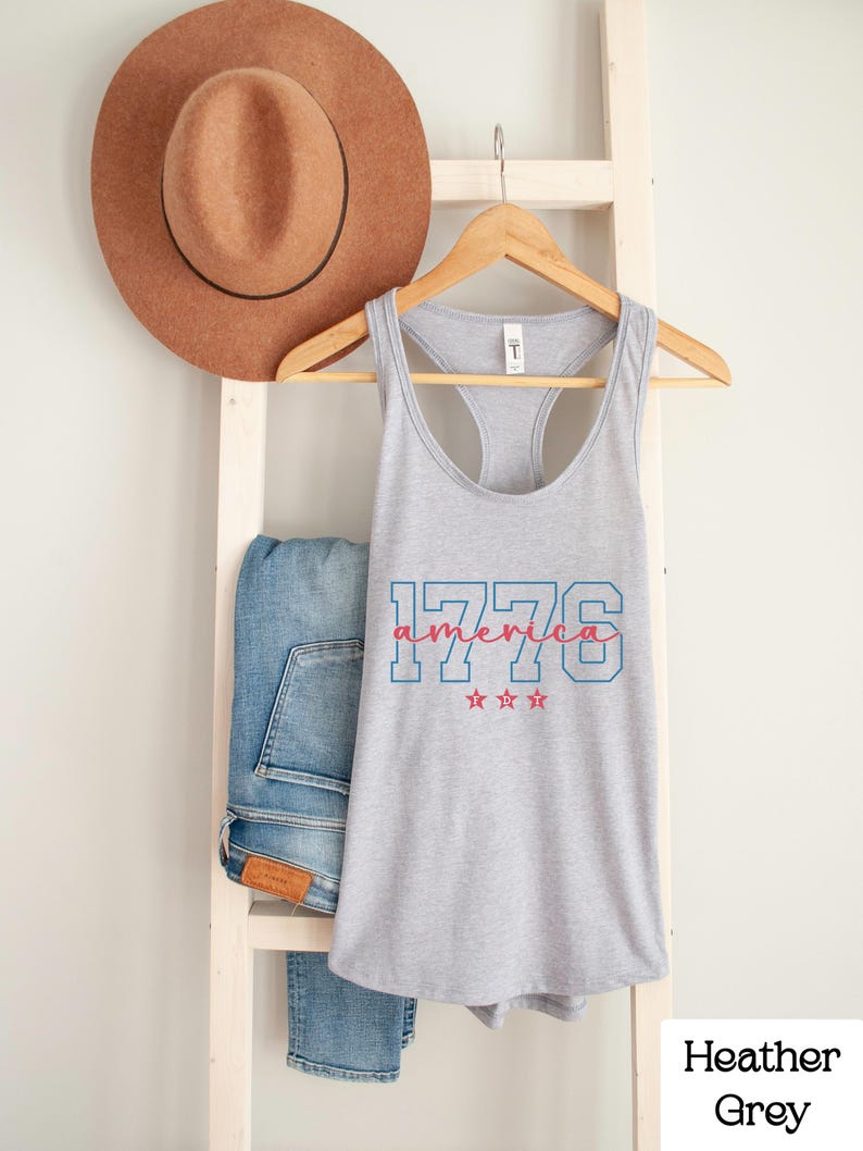 May include: A heather grey vest top with the text "1776 America" in red and blue. The top is displayed on a wooden hanger, with a brown hat and blue jeans in the background.