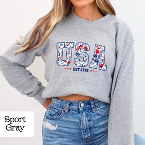 May include: A light gray sweatshirt with the word "USA" in a floral design, with the text "EST. 1776" below. The sweatshirt is paired with blue jeans.