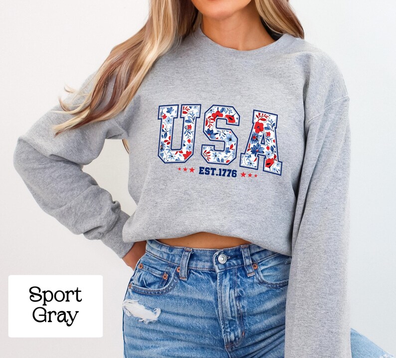 May include: A light gray sweatshirt with the word "USA" in a floral design, with the text "EST. 1776" below. The sweatshirt is paired with blue jeans.