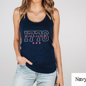 May include: A navy blue vest top featuring the text "1776 america" in white and pink. The design includes three pink stars. The top is a casual style.