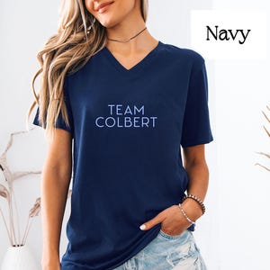 May include: Navy blue V-neck t-shirt with the text "TEAM COLBERT" in light blue. The shirt is a solid color and has short sleeves. The word "Navy" is in the upper right corner.