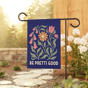May include: A navy blue garden flag with a floral design featuring pink and orange flowers and green leaves. The text "BE PRETTI GOOD" is at the bottom. The flag is displayed on a black metal stand in a garden setting.
