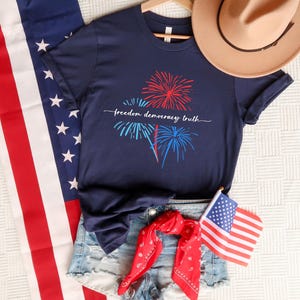 Puede incluir: Una camiseta azul marino de manga corta con un gráfico de fuegos artificiales rojos y azules y el texto blanco "freedom democracy truth" en el centro, junto con pantalones cortos de mezclilla azul claro, un sombrero beige y una bandera estadounidense.