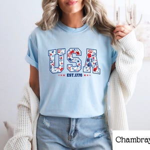 May include: Light blue t-shirt with the word "USA" in a floral design, with red, white, and blue flowers. The text "EST. 1776" is below the word "USA". The shirt is a casual style.