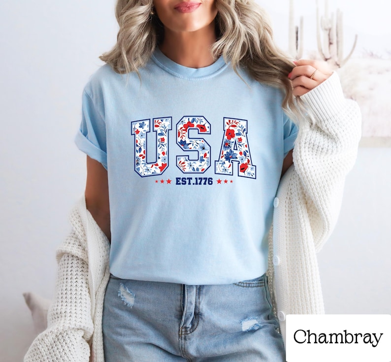 May include: Light blue t-shirt with the word "USA" in a floral design, with red, white, and blue flowers. The text "EST. 1776" is below the word "USA". The shirt is a casual style.