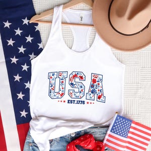 May include: White tank top with the word "USA" in a floral design, with the text "EST.1776". The tank top is displayed with a US flag, a hat, and denim shorts, evoking a patriotic theme.