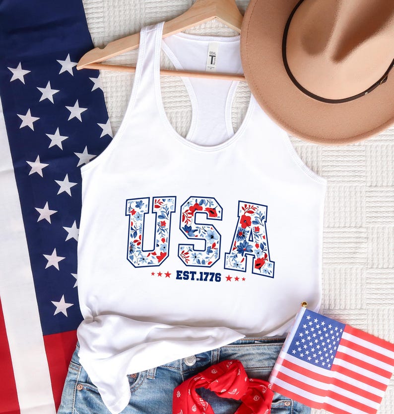 May include: White tank top with the word "USA" in a floral design, with the text "EST.1776". The tank top is displayed with a US flag, a hat, and denim shorts, evoking a patriotic theme.