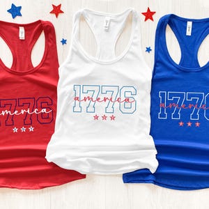 May include: Three tank tops in red, white, and blue. Each top has the number "1776" in a block font, with the word "america" in a script font underneath. Three small stars are below the text on each tank top.