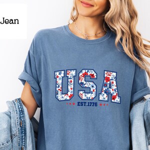 May include: A blue jean-colored t-shirt with the word "USA" in a floral design. The letters are white with a floral pattern of red and blue flowers. Below the word "USA" is "EST.1776" with red stars.