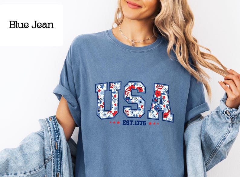 May include: A blue jean-colored t-shirt with the word "USA" in a floral design. The letters are white with a floral pattern of red and blue flowers. Below the word "USA" is "EST.1776" with red stars.