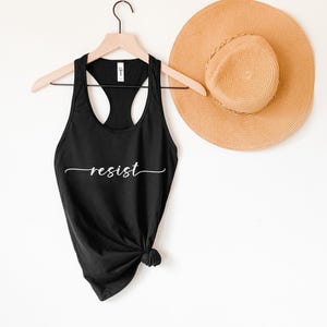 May include: A black tank top with the word "resist" written in white cursive script. The tank top is hanging on a hanger with a straw hat in the background.
