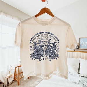 Könnte beinhalten: Ein cremefarbenes Kurzarm-T-Shirt mit einem dunkelblauen Grafikdruck von vier Hexen mit spitzen Hüten, die im Kreis tanzen. Der Text "GRAB HIM BY THE MIDTERMS" ist bogenförmig darüber angeordnet, umgeben von Sternen, Monden und Blattmotiven.