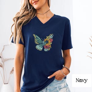 Può includere: Una maglietta blu navy a collo a V con una grafica di farfalla colorata fatta di fiori. La farfalla è centrata sulla maglietta.