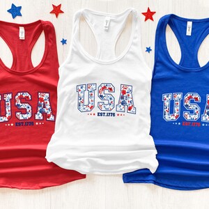 May include: Three tank tops in red, white, and blue. Each top features the word "USA" in a floral design, with "EST. 1776" below. Patriotic stars decorate the background, creating a festive display.
