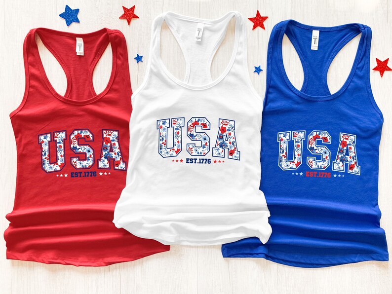 May include: Three tank tops in red, white, and blue. Each top features the word "USA" in a floral design, with "EST. 1776" below. Patriotic stars decorate the background, creating a festive display.
