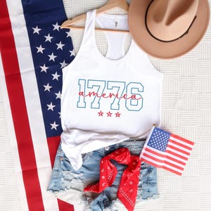 May include: A white vest top with the text "1776 America" in teal and red, alongside denim shorts, a red bandana, and a small American flag. A brown hat is positioned above the top, and an American flag is draped on the left.