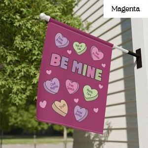 May include: A magenta flag with the words "BE MINE" and heart-shaped candies with phrases like "Free Press" and "Civil Rights." The flag is hanging outside on a white pole.